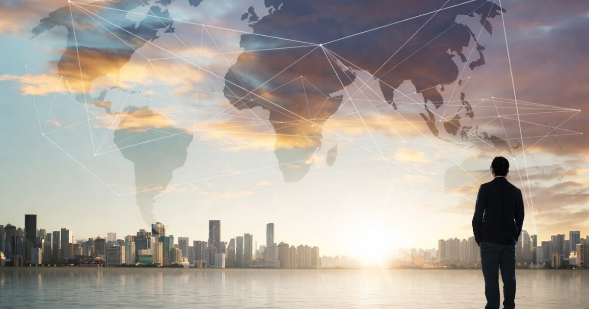 水辺に立つスーツ姿の成功者と見られる人物が夕暮れの都市を見つめ、その上空に世界地図と各地を結ぶネットワークの線が重ねて表現されている様子が写っている写真